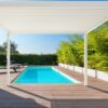 Ein moderner Außenpoolbereich verfügt über ein Holzdeck und eine elegante weiße Pergola mit Lamellendach für Schatten. Die Ränder des Decks sind mit Grünpflanzen gesäumt und in der Nähe steht ein Liegestuhl. Der klare blaue Himmel deutet auf einen sonnigen Tag hin.