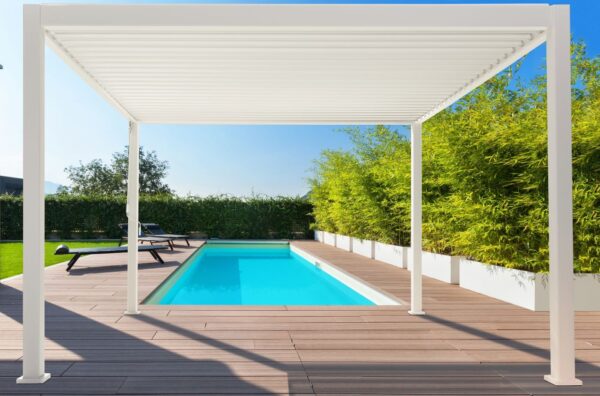 Ein moderner Außenpoolbereich verfügt über ein Holzdeck und eine elegante weiße Pergola mit Lamellendach für Schatten. Die Ränder des Decks sind mit Grünpflanzen gesäumt und in der Nähe steht ein Liegestuhl. Der klare blaue Himmel deutet auf einen sonnigen Tag hin.