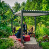 Eine Gruppe von Menschen unterhält sich unter einer manuellen Pergola mit Lamellendach in einem üppigen Garten. Sie sitzen um einen mit einer roten Tischdecke bedeckten Tisch inmitten von grünem Laub. Eine Person steht, während eine andere auf einem Korbstuhl sitzt. Auf dem Boden steht eine Laterne.
