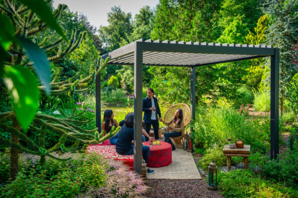 Eine Gruppe von Menschen unterhält sich unter einer manuellen Pergola mit Lamellendach in einem üppigen Garten. Sie sitzen um einen mit einer roten Tischdecke bedeckten Tisch inmitten von grünem Laub. Eine Person steht, während eine andere auf einem Korbstuhl sitzt. Auf dem Boden steht eine Laterne.