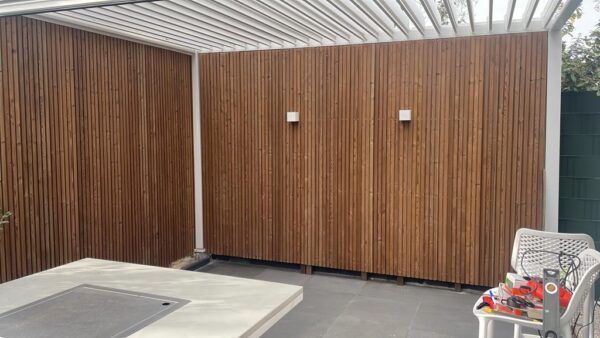 Die Außenterrasse punktet mit einem Lamellendach aus Holzwand und zwei weißen quadratischen Lampen darüber. Daneben stehen ein weißer Netzstuhl und ein Tisch mit verschiedenen Werkzeugen und Materialien, ergänzt durch einen dunklen Steinfliesenboden.
