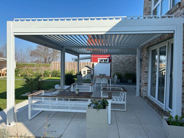 Die Terrasse verfügt über ein Lamellendach nach Maß (Preis auf Anfrage) mit modernen Möbeln wie Sofa und Esstisch. Im Hintergrund befindet sich ein kleines Spielhaus inmitten von Grün und einem Backsteingebäude, alles unter einem klaren blauen Himmel.