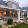Ein modernes Backsteinhaus mit einer geräumigen Terrasse verfügt über eine weiße Pergola mit Lamellendach und elektrischem Antrieb. Darunter befindet sich der Außenbereich mit einem Esstisch und Stühlen. Der Garten ist mit Steinen, Sträuchern und Zierpflanzen angelegt.