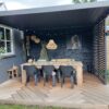 Ein gemütlicher Essbereich im Freien verfügt über einen Holztisch und schwarze Stühle unter einer eleganten Pergola mit Lamellen. Zur Einrichtung gehören eine große Hängelampe aus Korbgeflecht, Wandpflanzen und gerahmte Kunstwerke. Der geometrisch gemusterte Boden wird für zusätzlichen Charme von einer stilvollen Lamellenschiebewand oder einem festen Lamellenpaneel eingefasst.