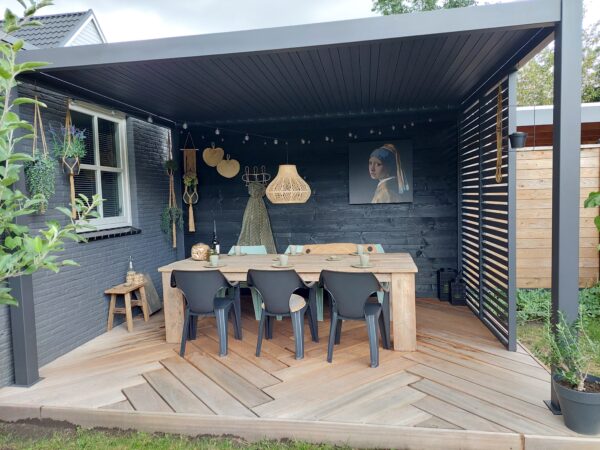 Ein gemütlicher Essbereich im Freien verfügt über einen Holztisch und schwarze Stühle unter einer eleganten Pergola mit Lamellen. Zur Einrichtung gehören eine große Hängelampe aus Korbgeflecht, Wandpflanzen und gerahmte Kunstwerke. Der geometrisch gemusterte Boden wird für zusätzlichen Charme von einer stilvollen Lamellenschiebewand oder einem festen Lamellenpaneel eingefasst.