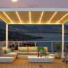 Eine moderne Dachterrasse mit einem maßgeschneiderten LED-Lamellendach nach Maß wirft in der Abenddämmerung ein warmes Licht. Entspannen Sie auf dem gemütlichen Sofa mit Kissen neben dem Couchtisch und Grill und genießen Sie den ruhigen Blick auf die Berge und den See bei Vollmond.