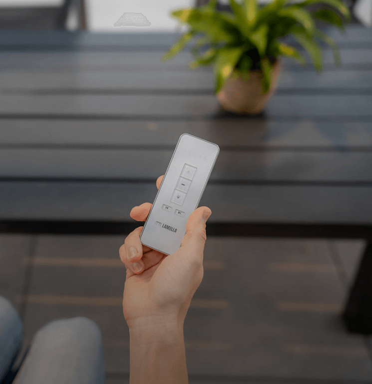 Une personne tient une télécommande à la main, la concentration intacte. À l'arrière-plan, une table design signée Über Lamella apparaît floue, accompagnée d'une plante verte en pot.