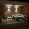 Eine gemütliche Außenterrasse bei Nacht mit sanfter Beleuchtung, mit einem L-förmigen Holzsofa mit Kissen und ein paar runden Tischen. Unter einer nachhaltigen Aluminiumüberdachung verbessern dekorative Pflanzen und strukturierte Wandkunst das Ambiente vor dem holzgetäfelten Hintergrund und schaffen eine warme und einladende Atmosphäre.