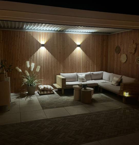 Une terrasse extérieure chaleureuse, baignée de lumière tamisée, accueille le soir un canapé en bois en L avec coussins et quelques tables rondes. Sous un auvent en aluminium durable, des plantes décoratives et des œuvres d'art murales texturées rehaussent l'ambiance sur fond de boiseries, créant une atmosphère chaleureuse et accueillante.