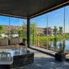 Ein moderner Wintergarten mit großen Glasfenstern und einer nachhaltigen Aluminiumüberdachung bietet einen Blick auf den Kanal. Im Inneren steht ein Korbsofa mit dunklen Kissen und einem passenden Couchtisch. Topfpflanzen zieren die Ecken, während draußen Gebäude und Bäume sichtbar sind.