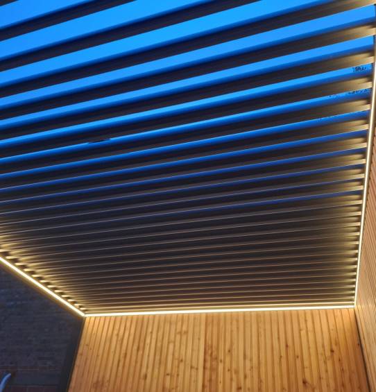 Un plafond moderne est composé de persiennes parallèles avec éclairage intégré, créant un motif rayé sur fond de ciel bleu. Le périmètre de la structure en aluminium durable est éclairé par des LED douces et chaleureuses qui mettent en valeur la texture des murs en bois et reflètent le style de Gaanderen.