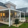 Ein modernes Backsteinhaus mit großen Fenstern und einer überdachten Terrasse mit Esstischen und Stühlen im Freien unter einer maßgeschneiderten Überdachung aus Aluminiumlamellen. Der Hof ist mit Steinen, Pflanzen und einem kleinen Trampolin angelegt. Der Himmel ist bedeckt.