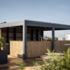 Eine moderne Outdoor-Dachbar mit Holzpaneelen und einem eleganten Metallrahmen bietet eine maßgeschneiderte Überdachung in Cromvoirt. Der Raum ist von Grünflächen unter einem klaren blauen Himmel umgeben, ergänzt durch Getränkehähne und hohe Tische mit Stühlen, die eine stilvolle und einladende Atmosphäre schaffen.