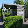 Eine moderne Pergola-Struktur mit horizontalen Lamellen ist von üppigen grünen Büschen umgeben. Zwei minimalistische Laternenpfähle stehen in der Nähe. Die Umgebung umfasst Bäume und einen Weg mit einer ruhigen, sonnigen Außenatmosphäre.