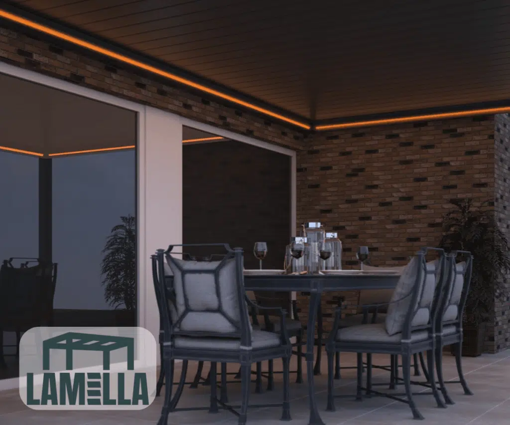 La salle à manger extérieure dispose d'une table avec six chaises et des verres. Une lumière orange discrète crée une ambiance chaleureuse. La scène est encadrée par un mur de briques arborant le logo « LAMELLA ». Un toit à lamelles réglable de multiples façons procure de l'ombre.