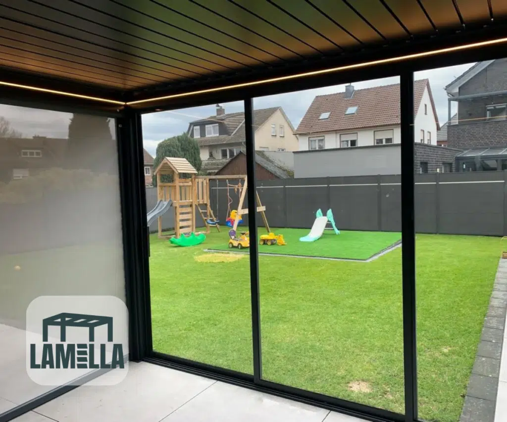 Depuis une terrasse moderne dotée d'élégantes portes coulissantes en verre à cadre noir et d'un toit à lamelles manuel (copie), vous pouvez profiter d'une vue imprenable sur une pelouse verdoyante avec une aire de jeux équipée de toboggans et de balançoires. La cour arrière clôturée offre une vue sur les maisons environnantes sous un ciel vaste et nuageux.