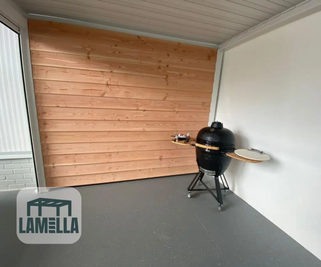 Un espace extérieur couvert avec un sol gris lisse et des murs clairs présente un barbecue en céramique noir sur roulettes avec une table d'appoint. Des panneaux verticaux en bois apportent une touche chaleureuse à l'arrière-plan. Le logo « LAMELLA », qui apporte une touche d'élégance, se trouve en bas à gauche et fait référence à l'élégant toit à lamelles réglable manuellement.