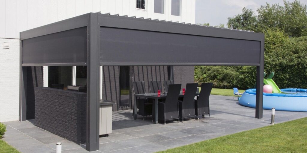 Une terrasse extérieure moderne avec une pergola grise et un socle en tuiles noires. En dessous se trouve une table à manger avec des chaises en osier noir posées sur des dalles en pierre. À l'arrière-plan, une piscine gonflable bleue pour enfants et une végétation luxuriante complètent le décor.