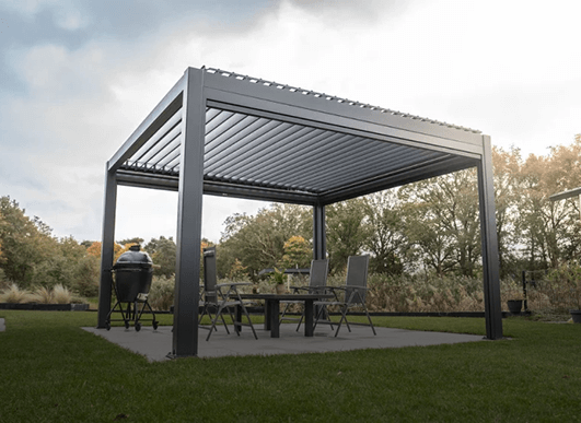 Une pergola métallique moderne avec un toit à lamelles se trouve sur une terrasse dans une cour arrière recouverte d'herbe et couvre une table avec quatre chaises et un barbecue. À l'arrière-plan, on aperçoit des arbres et des buissons sous un ciel partiellement nuageux.