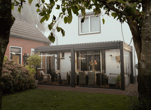 Une véranda moderne vitrée attenante à une maison blanche, avec des chaises et des tables en osier à l'intérieur, entourée d'espaces verts et d'arbres dans un jardin à l'arrière.