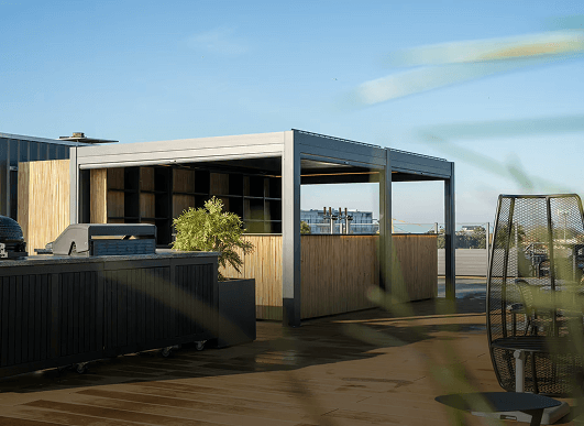 Une terrasse moderne sur le toit avec barbecue, cuisine extérieure, coin salon et pergola sous un ciel bleu clair avec quelques touches de verdure.